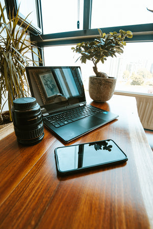 A bright window lite room shows a home office with laptop, cellphone and a camera lens.